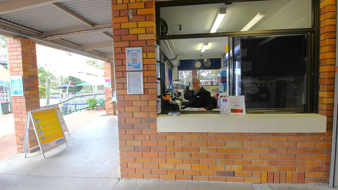 A closer look at the reception area. At the reception area's forefront, there are signs and frames associated with swimming. A receptionist is easily noticeable, wearing a smile as she attends to her tasks. Fluorescent lights cast their glow, and a wall clock hangs prominently within the reception. Additionally, a CCTV camera is positioned on the ceiling above the reception space.