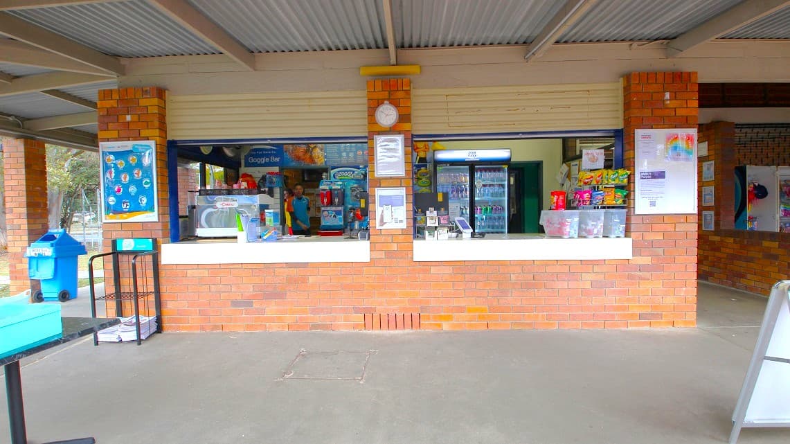 Frontal view of the reception area. The reception area's brick walls are adorned with posters and frames. An array of snacks and assorted refreshments are presented for the Aquatic Centre's visitors to see. Positioned to the left of the reception area is a blue waste bin. Beneath the poster, there is a black display stand in plain view.