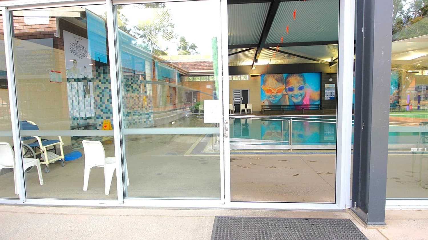 Glass doors with a view into an indoor swimming pool area. On the left, there are white plastic chairs and a wheelchair. The pool is visible through the glass, with a colourful mural on the far wall. The ceiling has a slanted design with lights, and the floor inside is tiled. A sign is posted on the door.