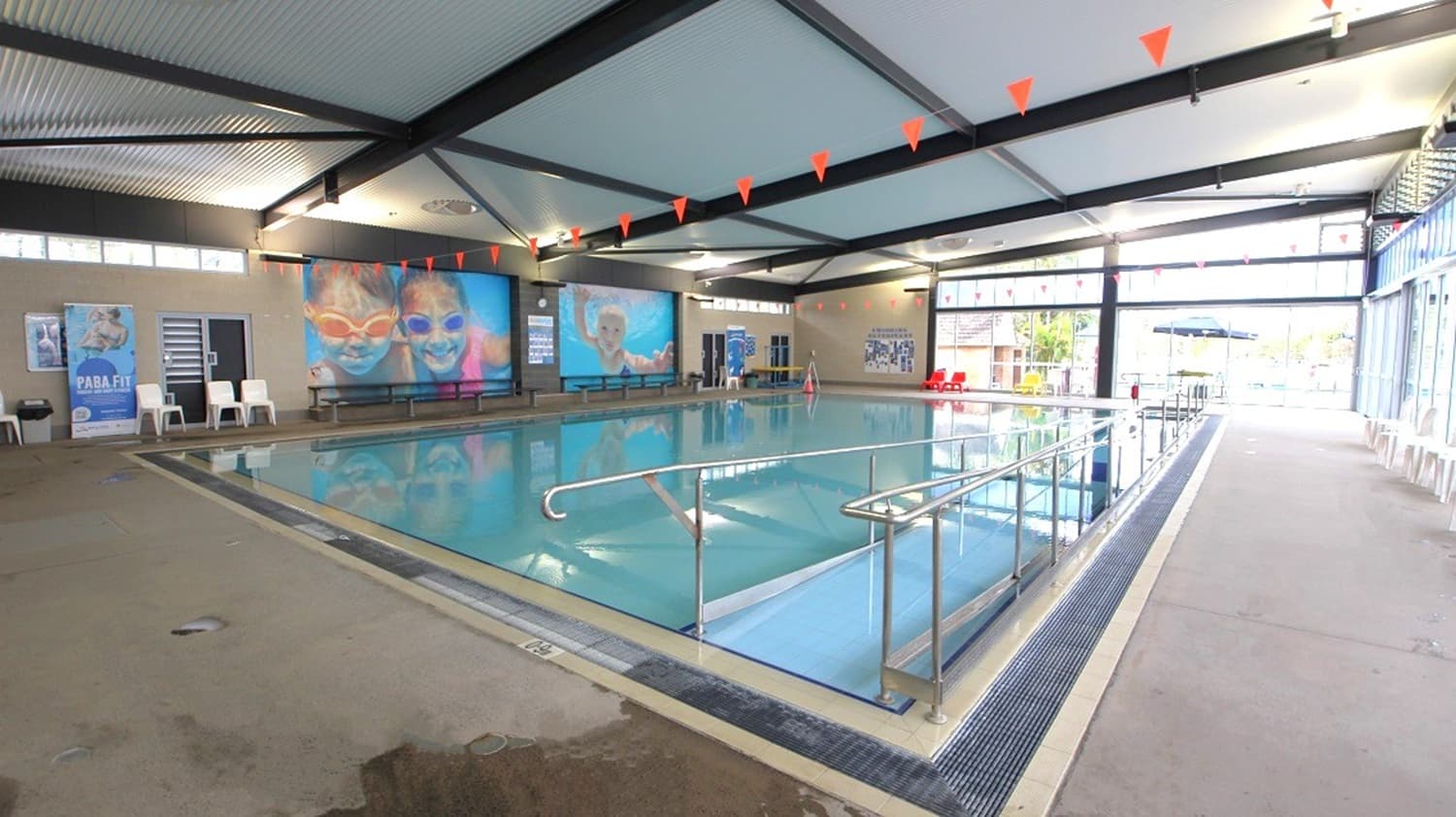Indoor swimming pool with a metal handrail leading into the water. The ceiling has exposed beams and red triangular flags hanging across. On the left, there are large posters on the wall and a few white chairs. The right side features a row of windows allowing natural light in, with more chairs along the wall. The floor is tiled, and the pool area is surrounded by a drainage grate. Outside, through the windows, some outdoor seating and umbrellas are visible.