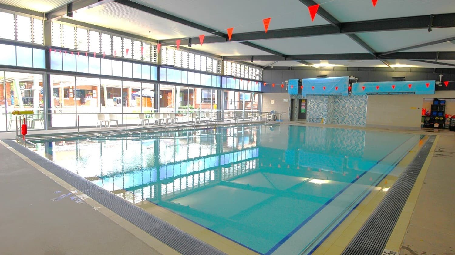 Indoor swimming pool with clear water, surrounded by a tiled deck. Large windows on the left let in natural light, with a view of an outdoor area. Red triangular flags hang above the pool. The ceiling is supported by dark beams, and the walls are light-coloured. On the right, there is a tiled wall and some pool equipment. The floor is tiled with drainage grates along the pool's edge.