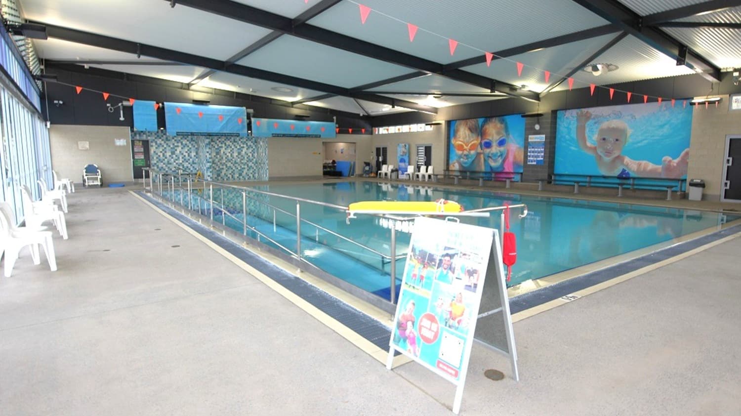 Indoor swimming pool with a tiled floor and a ceiling supported by dark beams. On the left, white plastic chairs line the poolside. In the centre, a metal railing separates the walkway from the pool, with a yellow float resting on it. A colourful signboard stands in the foreground. The right wall features large images of children swimming. Red triangular flags hang above the pool.