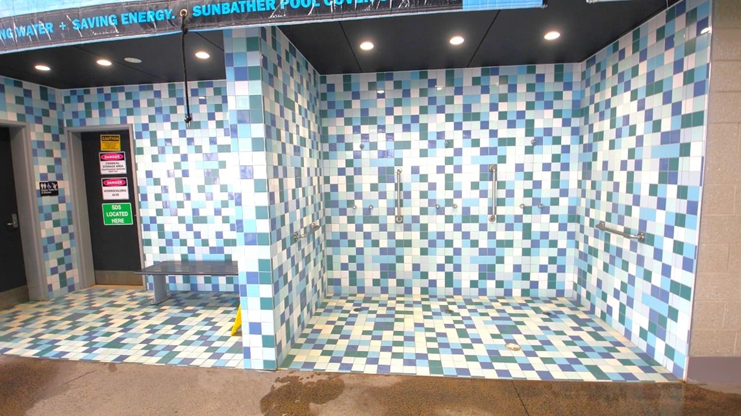 A tiled area with a mosaic pattern in shades of blue, green, and white. On the left, there is a doorway with signs and a small bench. The right side features a shower area with metal grab bars. The ceiling is black with recessed lighting. The floor continues the mosaic pattern, and there is a wet patch on the concrete floor in the foreground.