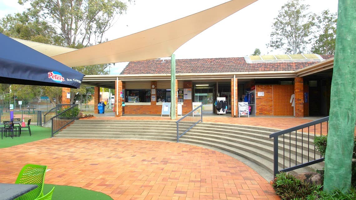 A brick building with a tiled roof is in the centre, featuring a shaded entrance with a few posters and signs. In front, there are curved steps leading down to a paved area. On the left, a large umbrella with "Peters" branding covers a seating area with green chairs and tables. A triangular shade sail is above the steps. Trees are visible in the background, and a green pole supports the shade sail.