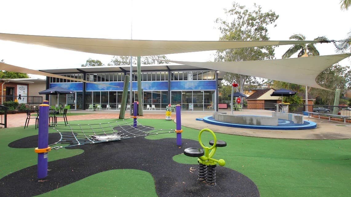 A playground with a green and black rubberised surface. On the left, there's a climbing net structure supported by purple and orange posts. In the centre, a spring-mounted seat with a green handle. On the right, a circular platform with a railing. Above, large white shade sails are stretched across. In the background, a two-storey building with large windows and a blue facade. Trees and additional structures are visible beyond the building.