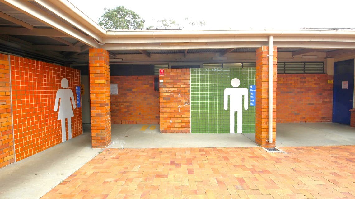A public restroom facility with two entrances. On the left, the entrance is marked with a white female figure on orange tiles. On the right, a white male figure is displayed on green tiles. The structure is made of brick with a tiled floor. The ceiling is covered with a corrugated roof. There are trees visible in the background above the roofline.