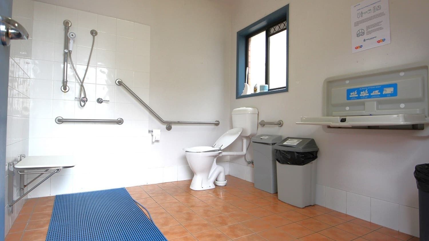 Accessible bathroom with a white toilet on the right, equipped with grab bars. A baby changing station is mounted on the wall above a bin. A window is above the toilet. On the left, a shower area with a foldable seat and more grab bars. The floor is tiled in a light brown colour, and a blue non-slip mat is placed near the shower. The walls are a mix of white tiles and light paint.