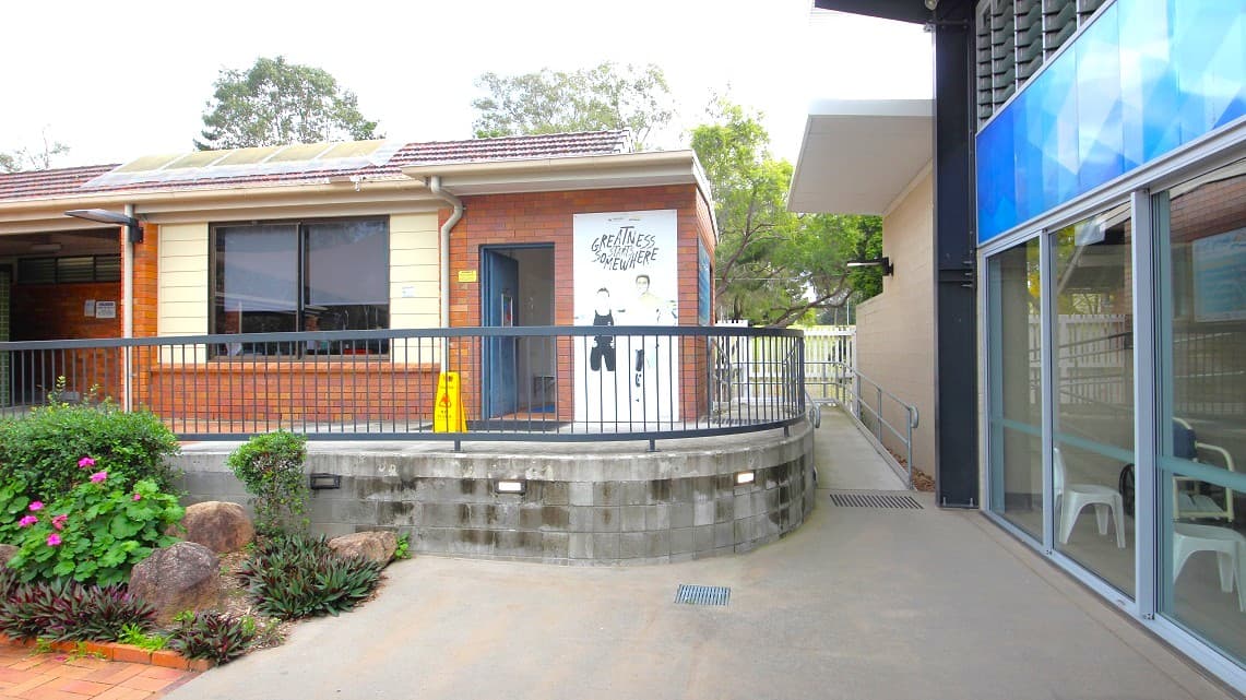On the left, a single-storey brick building with a tiled roof and a large window. A mural on the wall near the entrance features two figures and text. A yellow caution sign is visible near the door. In the foreground, a garden with rocks and plants. On the right, a modern building with large glass windows and a blue panel. A pathway runs between the two buildings, with a railing along the edge of the brick structure. Trees are visible in the background.