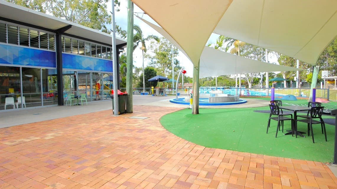 On the left, there is a modern building with large glass windows and a blue facade. In front of it, there are tables and chairs. The ground is paved with reddish-brown bricks. On the right, there is a green artificial turf area with a round table and chairs. A large shade sail is overhead, and beyond it, a pool area with water features and palm trees is visible. A metal fence encloses the pool area in the background.