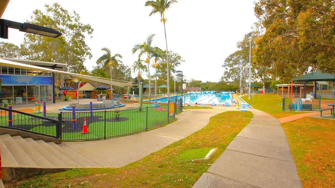 A swimming pool area with a fenced playground on the left, featuring a circular play structure and shade sails. Palm trees are scattered throughout the area. On the right, there is a large outdoor swimming pool with lanes, surrounded by a paved walkway and grassy areas. A few picnic tables and shelters are visible. The background is filled with tall trees, and the sky is overcast.