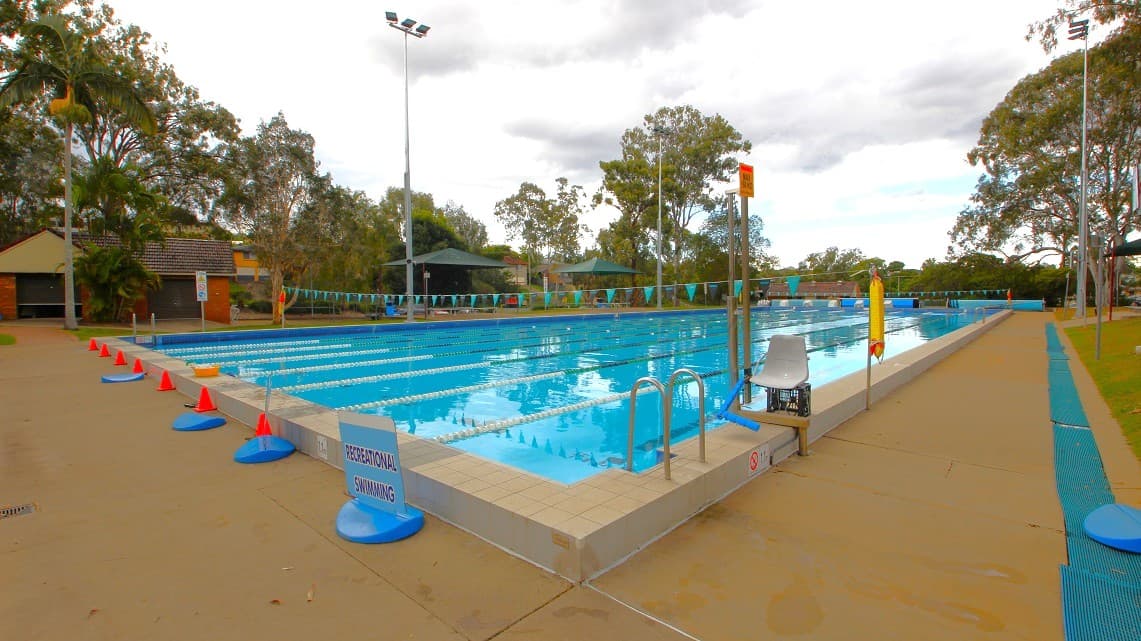 An outdoor swimming pool with multiple lanes, bordered by a concrete deck. On the left, there are red and blue cones along the pool's edge. A sign reads "RECREATIONAL SWIMMING." On the right, a metal ladder and a lifeguard chair are visible. Trees and a few buildings are in the background, with tall floodlights around the pool area. The sky is overcast.