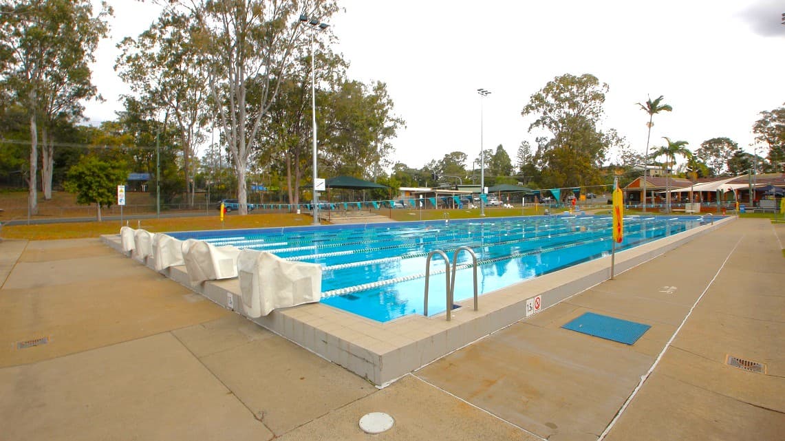 An outdoor swimming pool with multiple lanes, surrounded by a concrete deck. On the left, there are starting blocks and tall trees in the background. On the right, a row of palm trees and a covered seating area are visible. The pool has metal ladders for entry, and there are lane ropes dividing the swimming lanes. The area is well-lit with tall floodlights, and the sky is overcast.