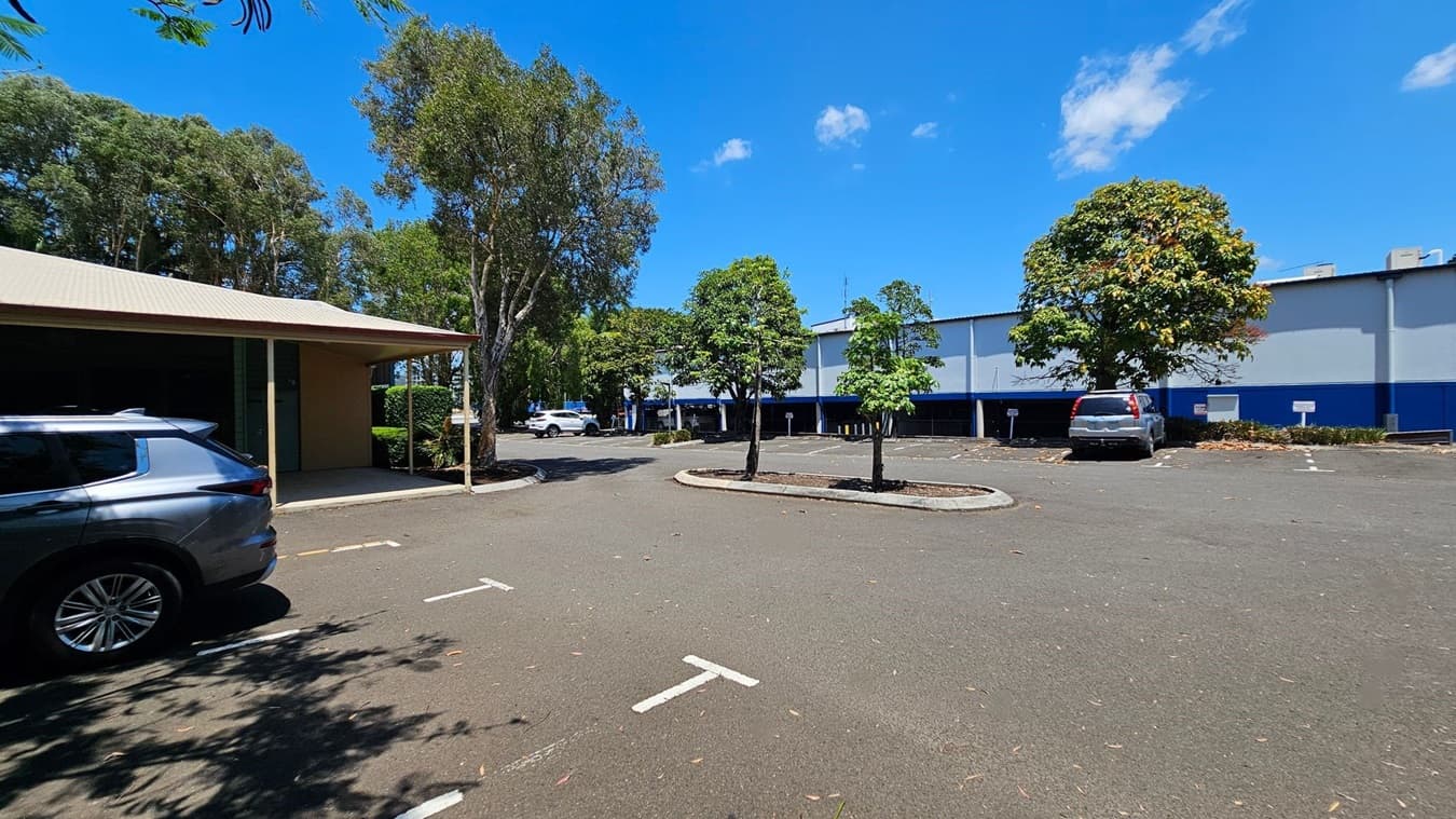 A car park with a few vehicles is visible. On the left, a grey car is parked near a building with a beige roof and green doors. In the centre, there are several trees planted in the parking area. On the right, a silver car is parked near a large building with a blue and white exterior. The ground is paved, and there are some leaves scattered around. Trees and greenery are in the background.