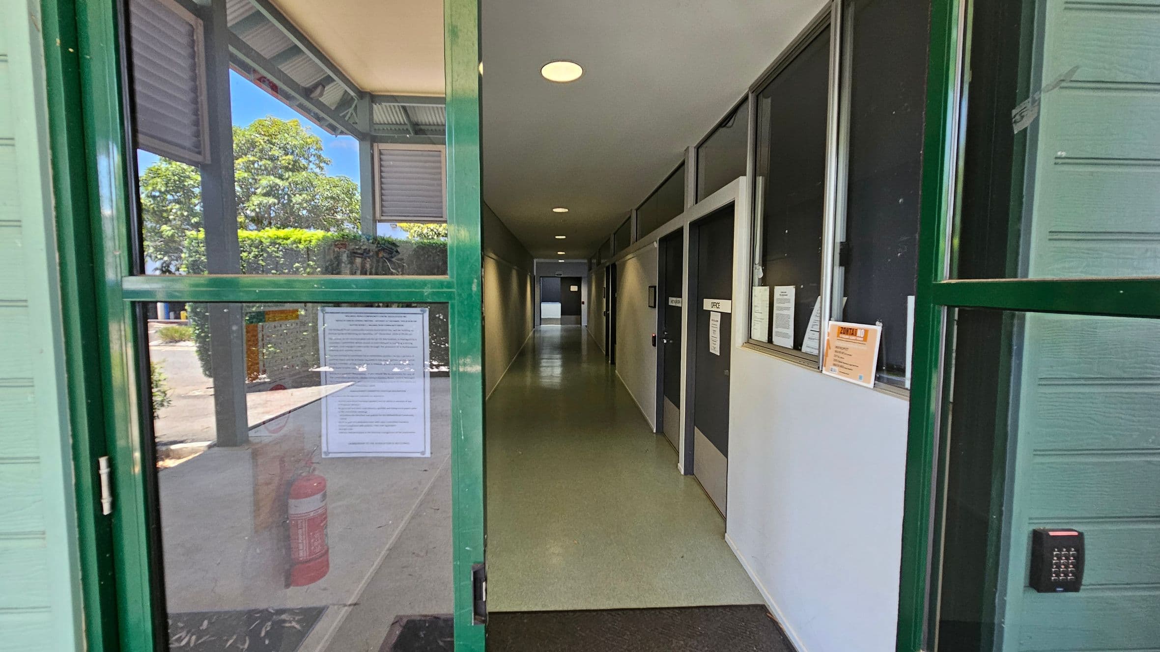 A set of green-framed glass doors, one open, leads into a building with light green wooden panelling. The floor outside is covered with a dark mat and scattered leaves. To the left, a beige wall displays a poster. Inside, a corridor with a green floor is visible. On the right, a small bush is next to a railing. A plaque is mounted on the wall above the bush.