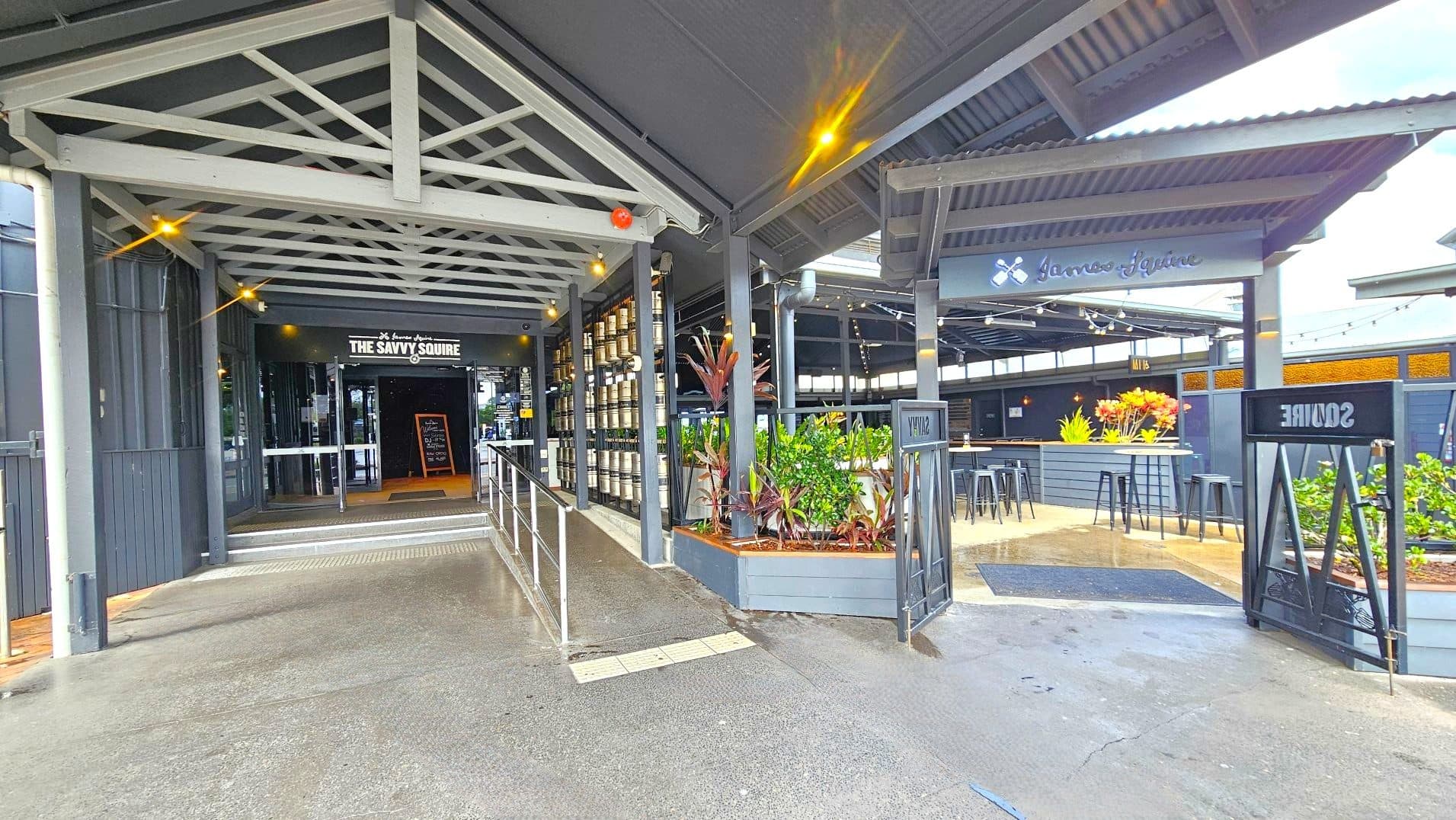 The image shows an entrance to a venue named 'The Savvy Squire'. The entrance features a covered walkway with a metal truss roof and a ramp with handrails leading to glass doors. To the right, there's an open seating area with high tables and stools, surrounded by planters with greenery. The flooring transitions from concrete to a textured surface at the entrance. Signage is visible above the entrance and seating area.