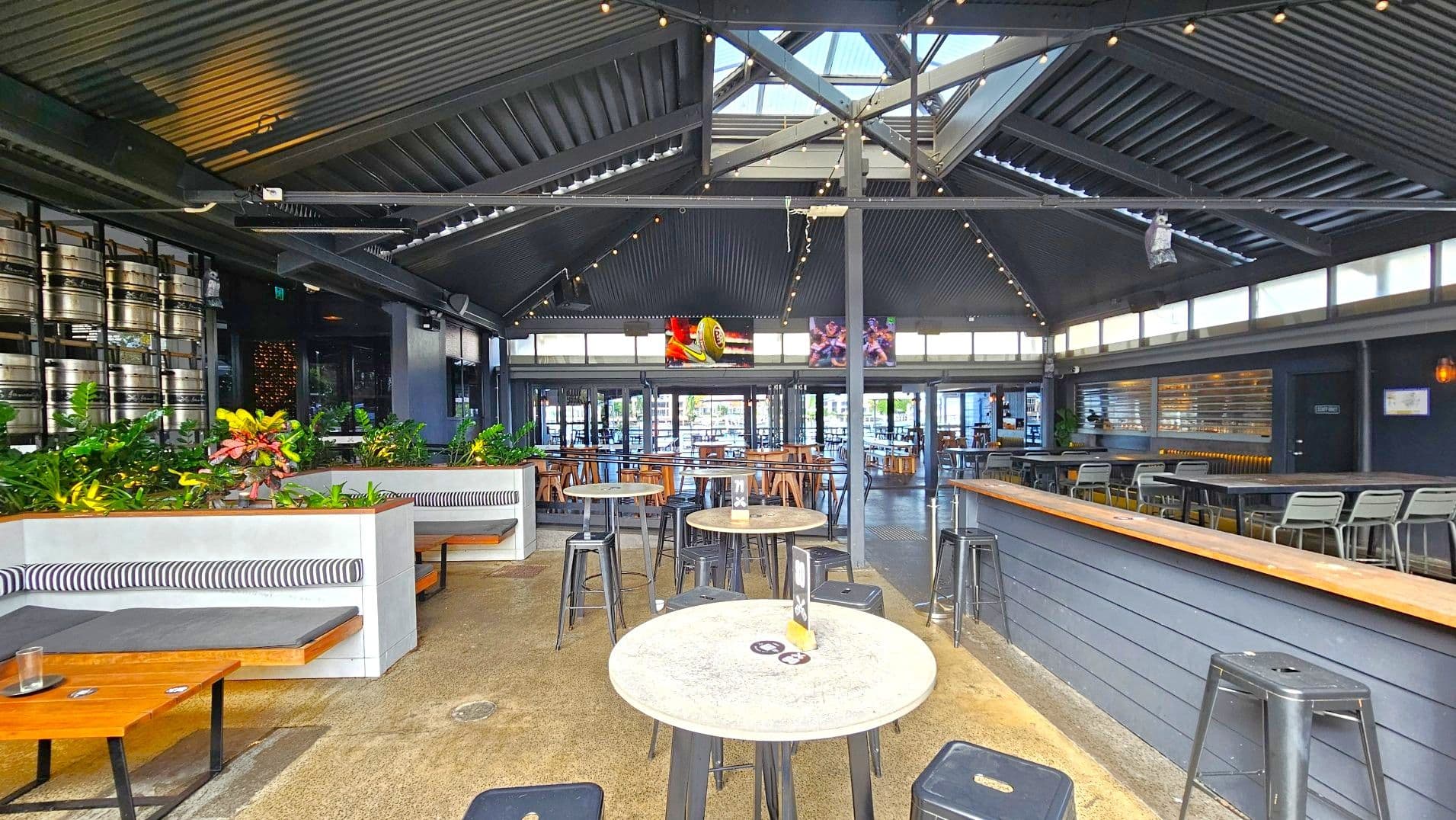 The interior of a modern bar with a high, dark corrugated metal ceiling featuring skylights. The space includes a mix of seating options: cushioned booths with striped backs, round tables with metal stools, and a long bar with high chairs. Green plants add a touch of nature. Large windows and glass doors line the far wall, allowing natural light. Kegs are stacked on one side, and two screens display sports images.
