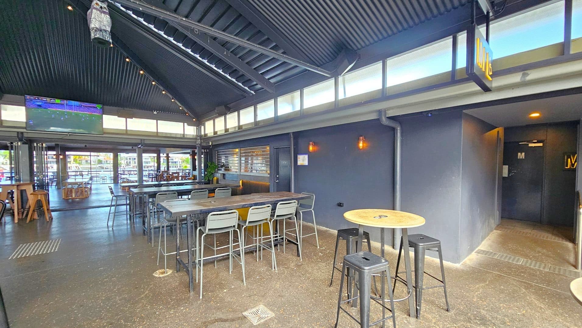 The interior of a modern bar with a high, corrugated metal ceiling and large windows near the roof allowing natural light. A large TV screen is mounted on the wall showing a sports game. The space features high tables with metal stools and a polished concrete floor. A sign marked 'M' indicates a restroom entrance on the right. The area feels open with visible seating extending outside.
