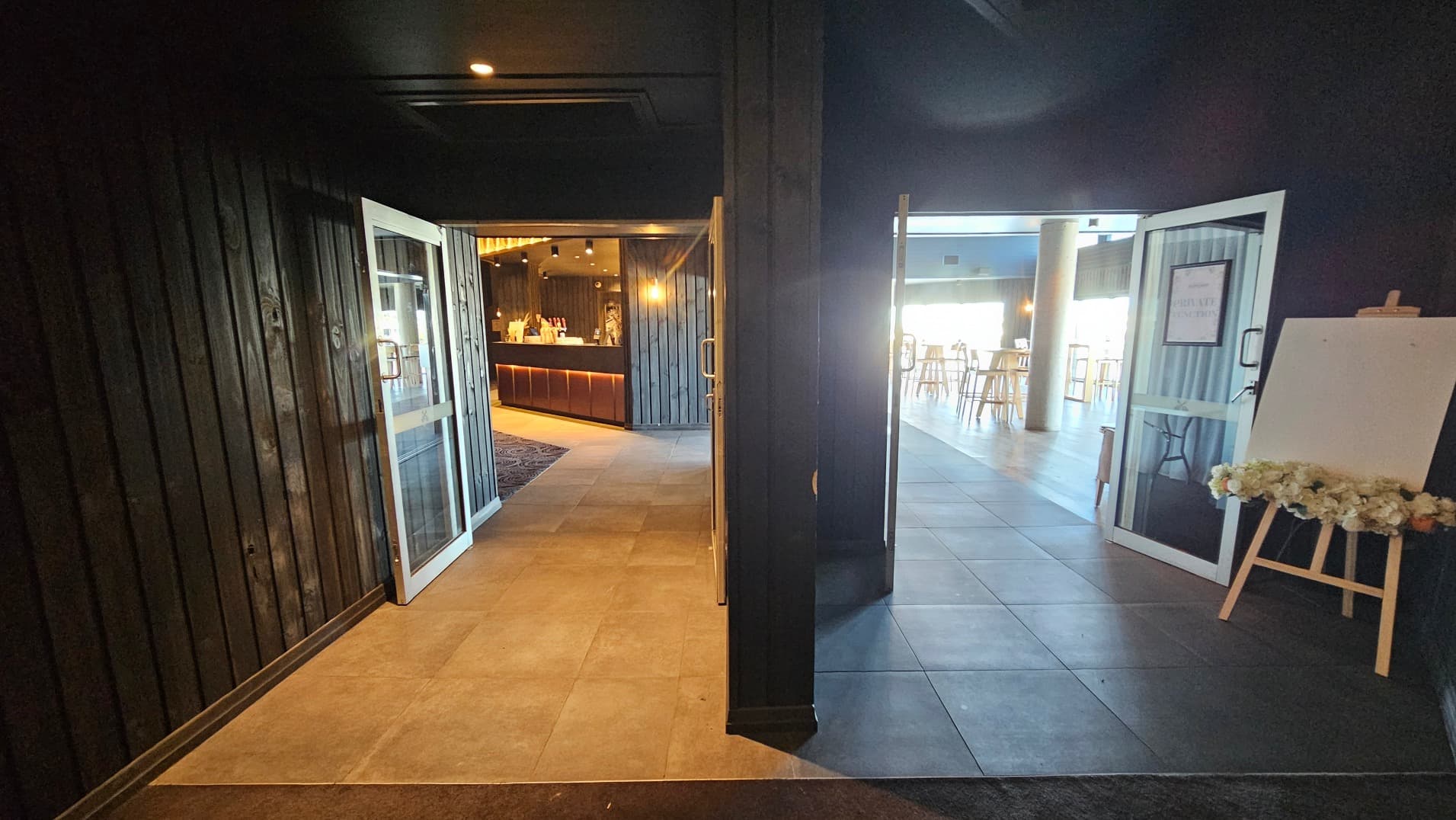 The image shows an interior space with two open doorways leading to different areas. On the left, a doorway opens to a warmly lit bar area with wooden panelling and a counter. On the right, another doorway leads to a brighter space with high tables and stools, featuring a large window. The floor transitions from dark tiles to lighter tiles. An easel with floral decorations stands near the right doorway.