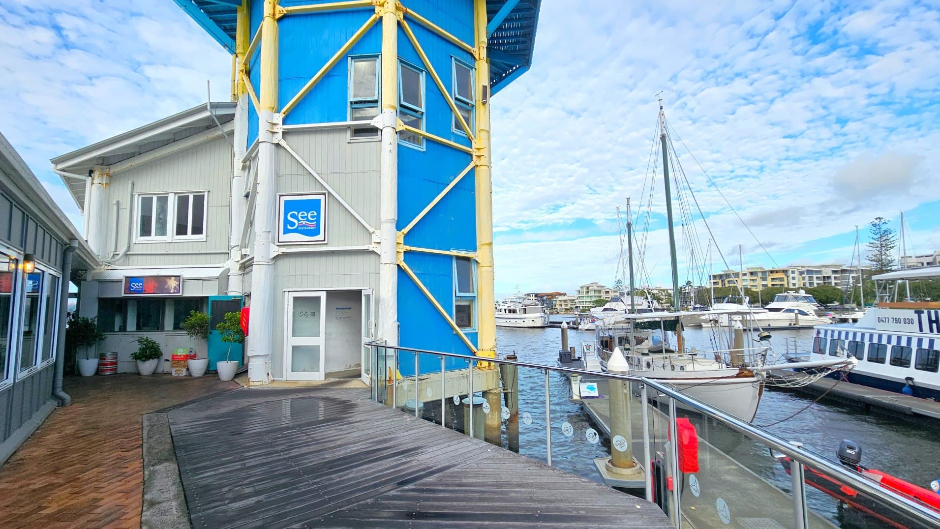 A waterfront scene featuring a two-storey building with a blue and grey facade, supported by yellow beams. The building has a sign reading 'See Restaurant'. A wooden boardwalk with a metal railing runs alongside, leading to a marina filled with various boats and yachts. Potted plants are placed near the entrance of the building. The area feels open with views of distant buildings across the water.