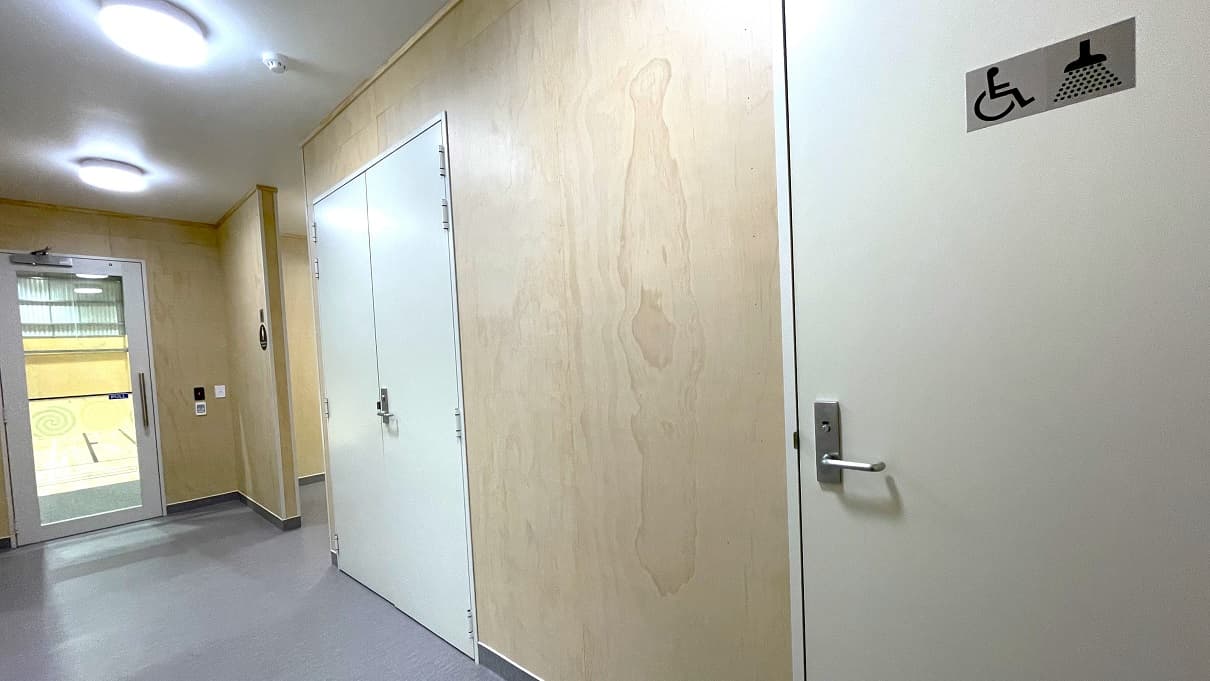Accessible bathroom and shower at the Gallagher Recreation Centre. The accessible bathroom has a disabled symbol and a shower head with water droplets on the off-white painted door. Before the accessible bathroom is a off-white painted double door and a hallway right after the glass entrance doors. All doors have a lever type, stainless steel door handle.