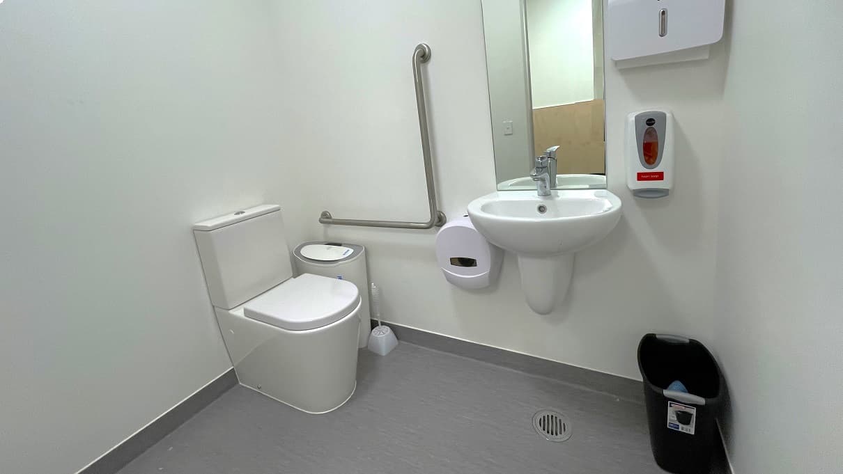 The bathroom has a grey floor and a white wall with dark brown painted bottom. On the left side of the toilet is a white/grey air purifier, a white toilet brush, and grab bars fixed to the white wall. There is also a low-set hand basin, a mirror, a liquid hand soap dispenser, and a toilet paper roll dispenser. A small black garbage bin is placed against the wall opposite the toilet.
