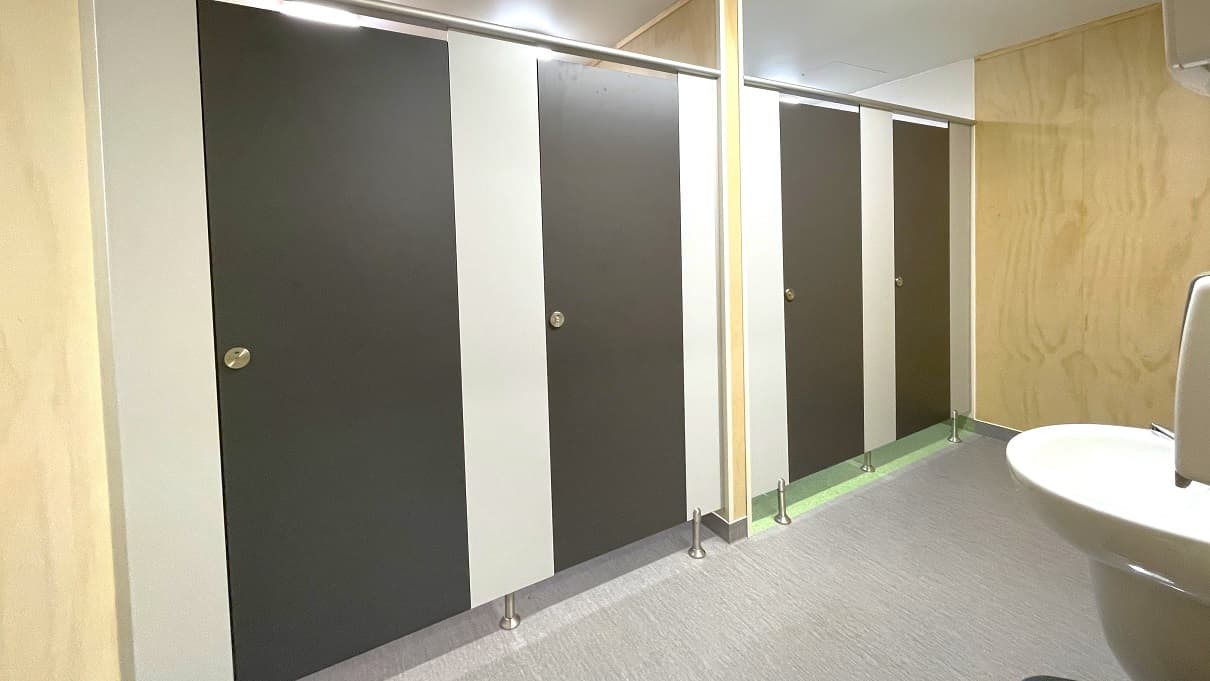 Other bathrooms in Gallagher Recreation Center. The bathroom has smooth, dark brown doors and small circular metal with green and red visual options to notify guests when occupied or not. A wall in the middle separates the bathroom stalls. The bathroom stalls have grey floor tiles, while the other two have light green floor tiles. Outside the bathroom stalls is a low-set hand basin with a liquid hand soap dispenser.