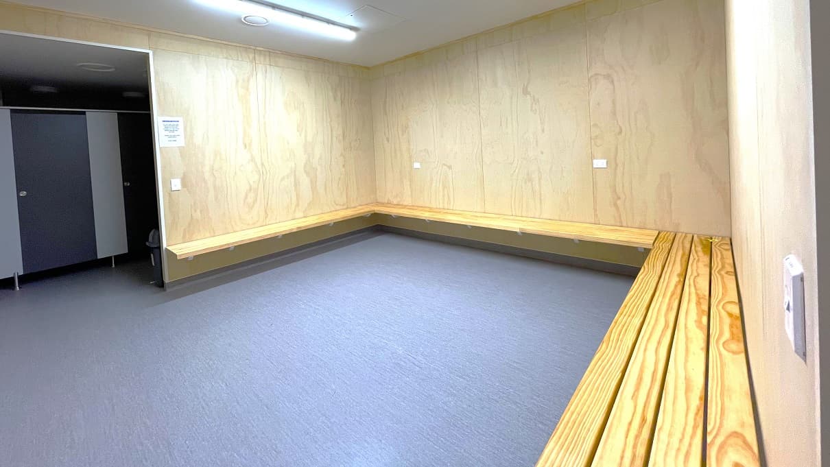 Seating area outside the bathroom stalls in Gallagher Recreation Center. Alongside the walls are four light brown wood planks that serve as seats. Similar to other areas, this has a brown wall and a white ceiling. On the right wall is the light switch. The area is illuminated by a fluorescent light. There is a small, black garbage bin in front of the bathroom stalls.