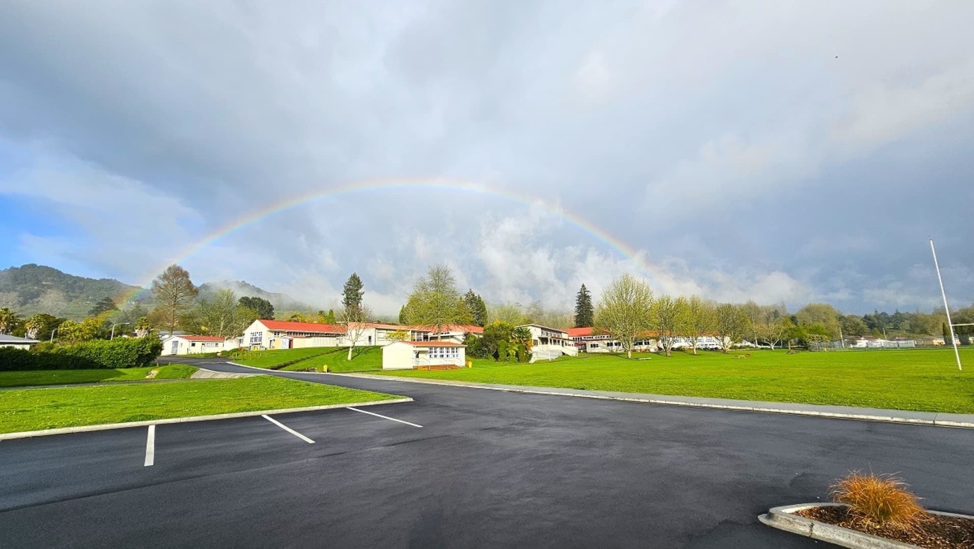 A paved parking area in the foreground with white lines marking spaces. To the right, a small patch of grass with a few plants. In the background, a series of white buildings with red roofs are surrounded by green lawns and trees. A faint rainbow arcs across the sky above the buildings. Hills and more trees are visible in the distance.