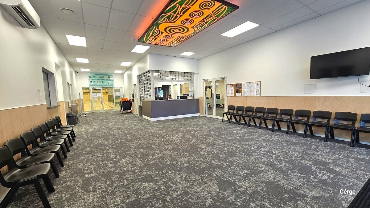 Grey carpet tiles flooring in the reception area. Black plastic chairs are placed against the brown-painted part of the wall on either side of the entrance. The ceiling is painted white with alternating rectangular lighting patterns and a green and yellow-orange Maori artwork. A TV is mounted on the right wall, and further beyond is a wooden bulletin board. To the right of the reception desk is the gym, to the left is the kitchen and bathrooms, while directly ahead is the stadium.