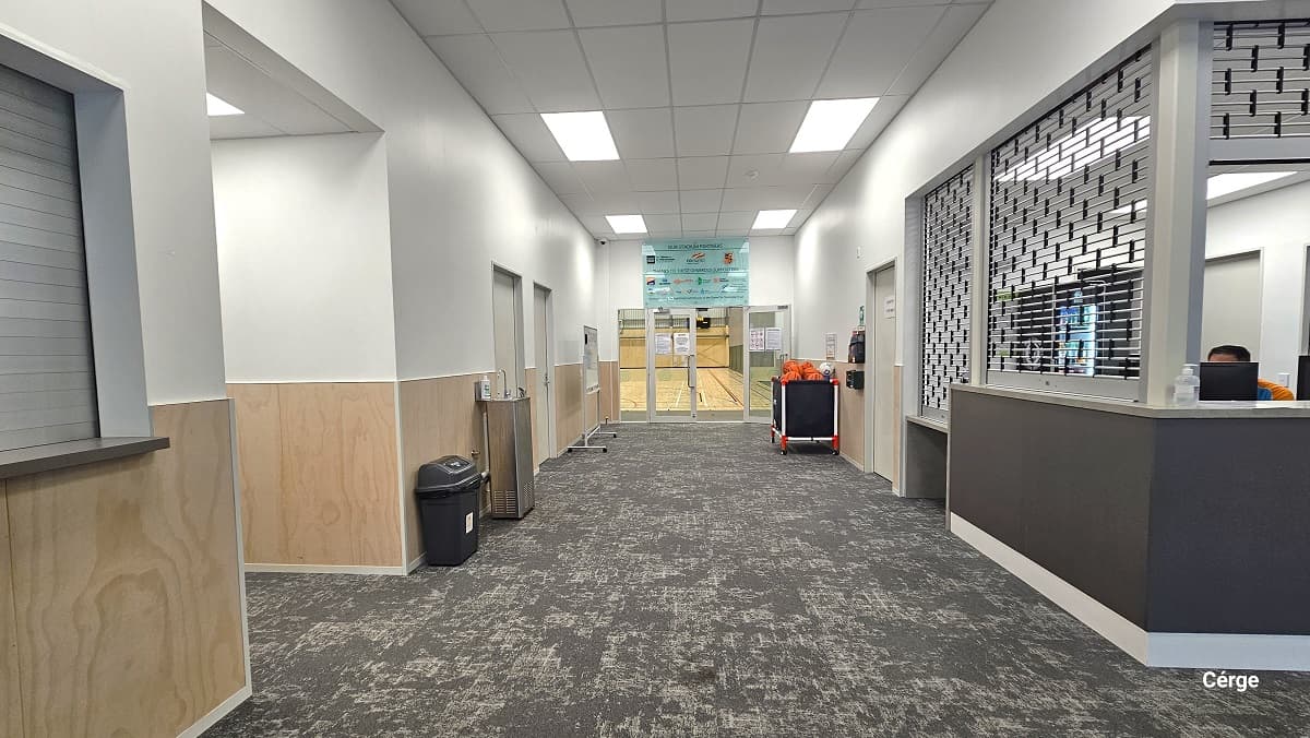 The reception desk at Gallagher Recreation Centre can be distinguished by the aluminum roller grille and a marble countertop with a grey-painted wall. There is another door next to the reception desk, while there are multiple doors opposite it. A small black garbage bin, a water fountain, and a mobile whiteboard can be found on the left side of the hallway leading to the stadium. On the right is a black storage ball rack. There is a light blue glass signboard atop the glass doors of the stadium.