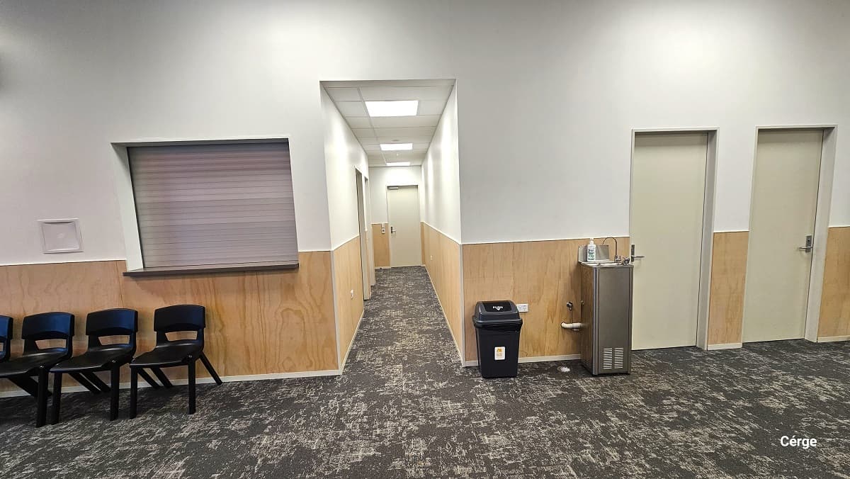 Kitchen and bathroom section at Gallagher Recreation Centre. On the left side, before the hallway, is a small window with an extended flat surface and several black plastic chairs. On the right side is the small black garbage bin, a water fountain, and two off-white painted doors. The image also shows a grey carpet tiles flooring and a combination of a brown wooden lower part and a white-painted upper wall and ceiling.