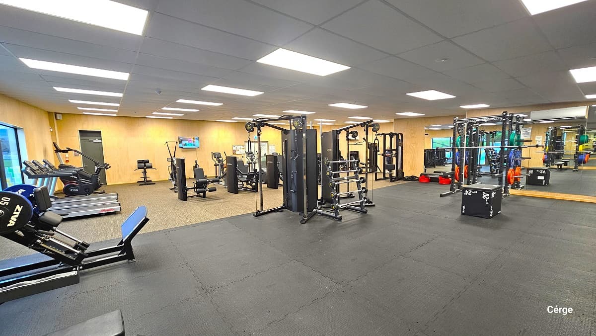 A closer look at the space inside the gymnasium near the TV mounted on a wall. The treadmills are aligned next to each other on the wall near the exit glass doors. This image also shows the large floor-to-ceiling mirror and an air conditioning unit inside the gymnasium on the right wall standing opposite the entrance doors. There is a large black box in the middle of the black gym mat floor.