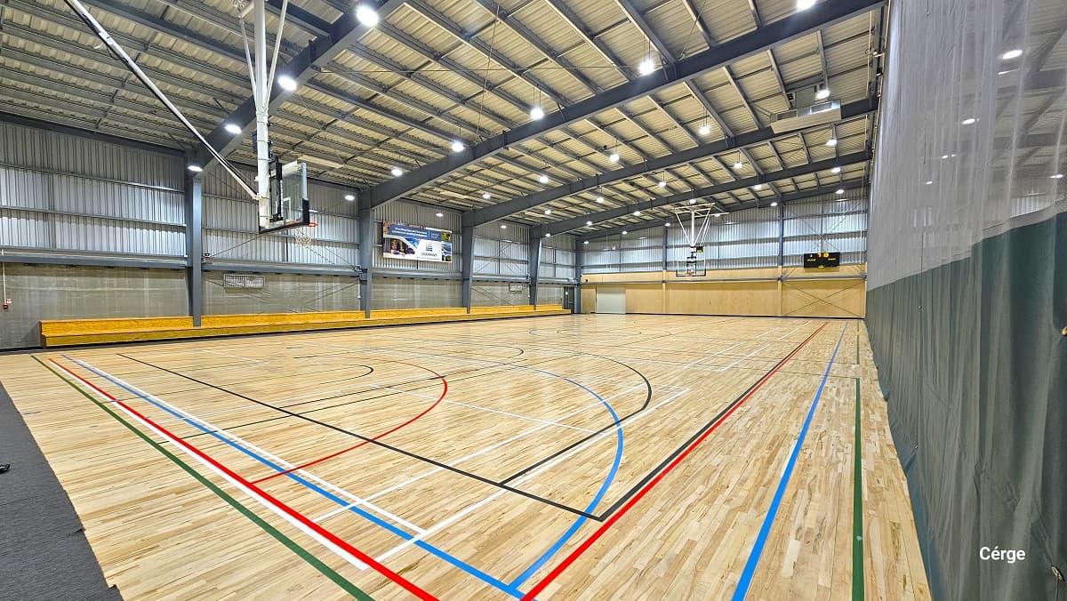 Side view of one of the basketball courts in the Sports stadium. The other court is separated by a curtain with a dark lower part. The floor surface changed from carpet tiles to timber floors with a highly polished finish which creates a low-level glare from the fluorescent lighting. The stadium has a very high ceiling made of off-white corrugated iron and wide grey steel beams. There is a two-tiered seating along two sides of the stadium.