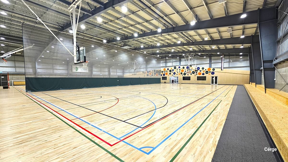 View of the basketball court standing in front of the two-tiered brown seating on the right side. There is a grey mat that separates the court from the seating area. Two glass doors at the back with white metal frames can be found within the brown-painted wall at the back with colorful circular designs. The other court has a low net wrapped around the red pole at the opposite ends.
