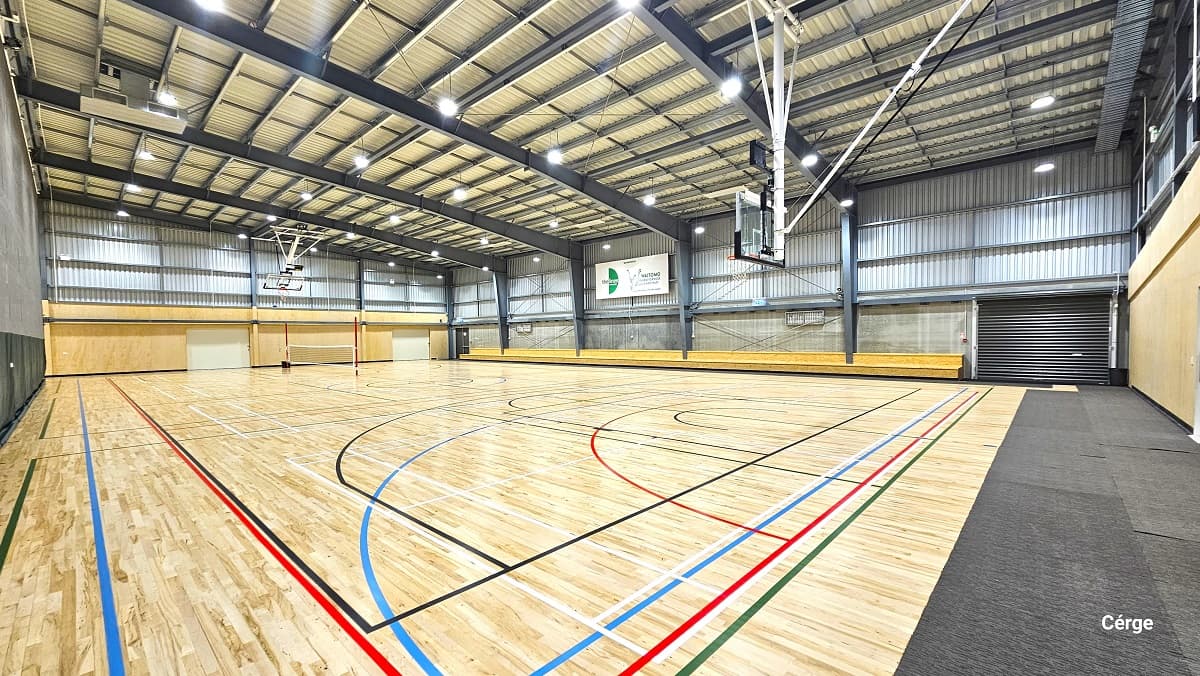 View of the second court with a low white/black wrapped around two red poles at opposite sides. The right end of the court is covered with grey mats, while the rest of the court has polished timber floors with red, black, blue, white, and green lines. There is also a two-tiered brown seating along the stadium side. On the upper part of the grey corrugated wall to the right is a large, white advertisement poster.