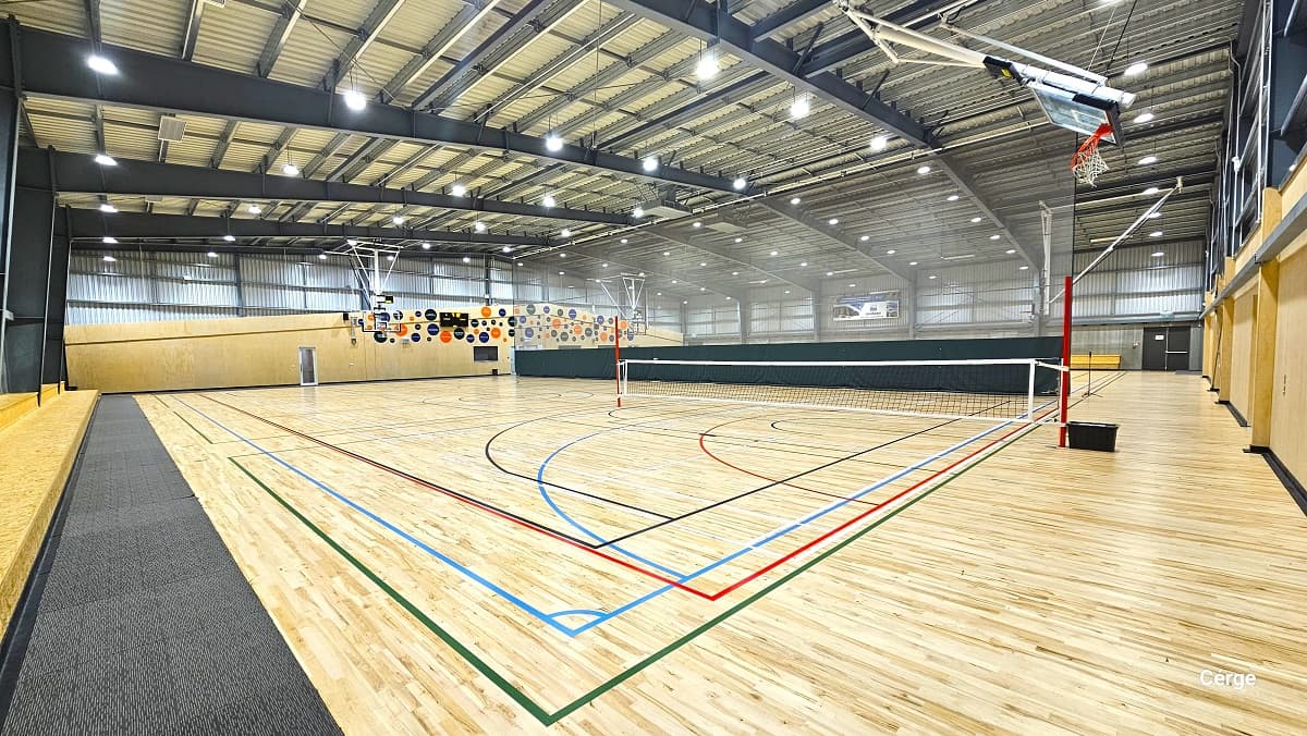 A closer look at the low net wrapped around two red poles placed at opposite ends. The basketball ring is slightly inclined towards the grey corrugated wall. On the left is a two-tiered seating alongside the stadium and grey mats. Also visible from this position are the colorful round designs on the wall above the glass entrance doors.