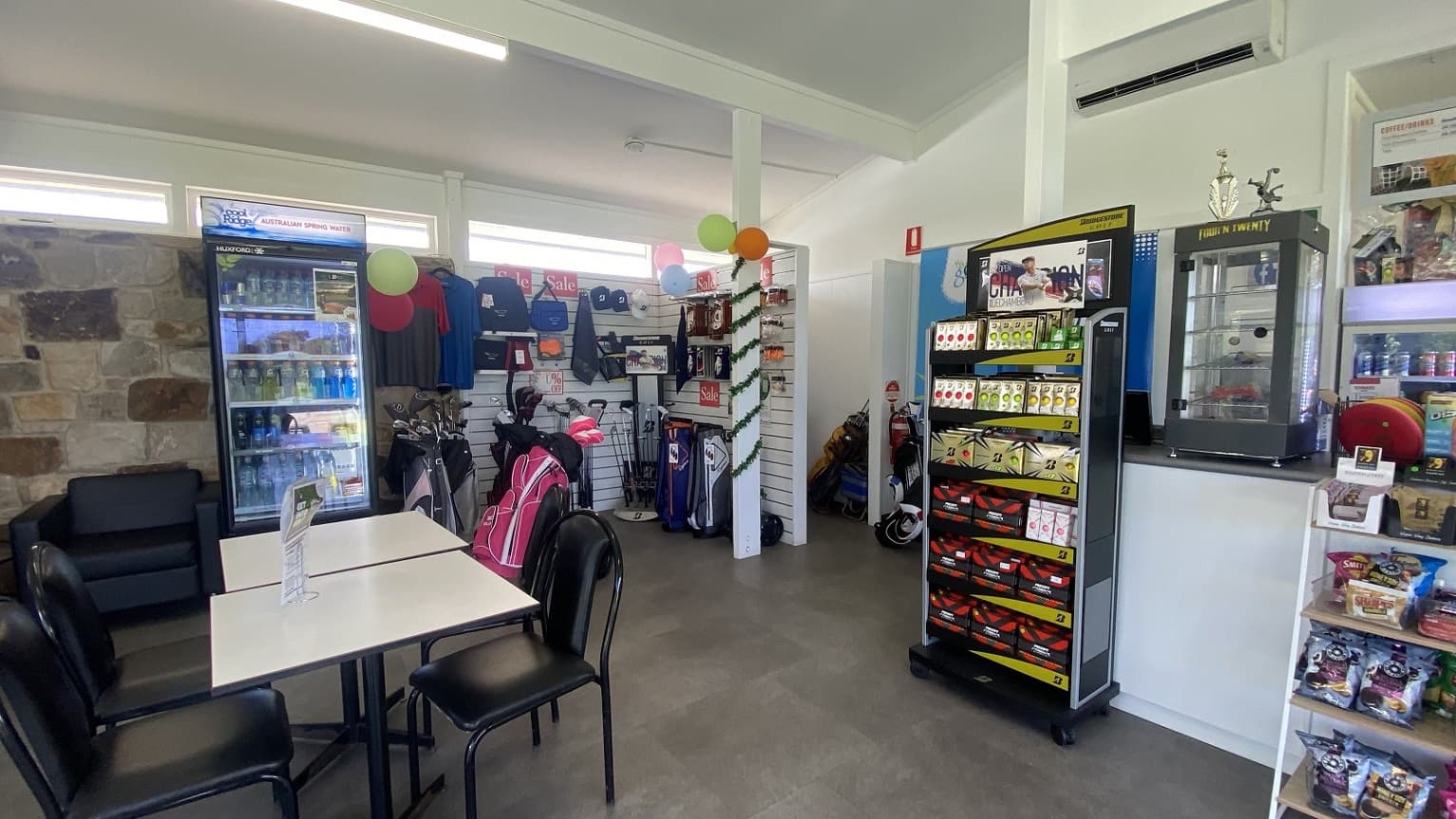 To the left of the countertop area is the retail space for golfing equipment, such as metal golf clubs, golf bags, shirts, golf caps, and more. Beside the golf retail space are a fridge and a black sofa. The retail area is decorated with colorful balloons and Christmas garlands.