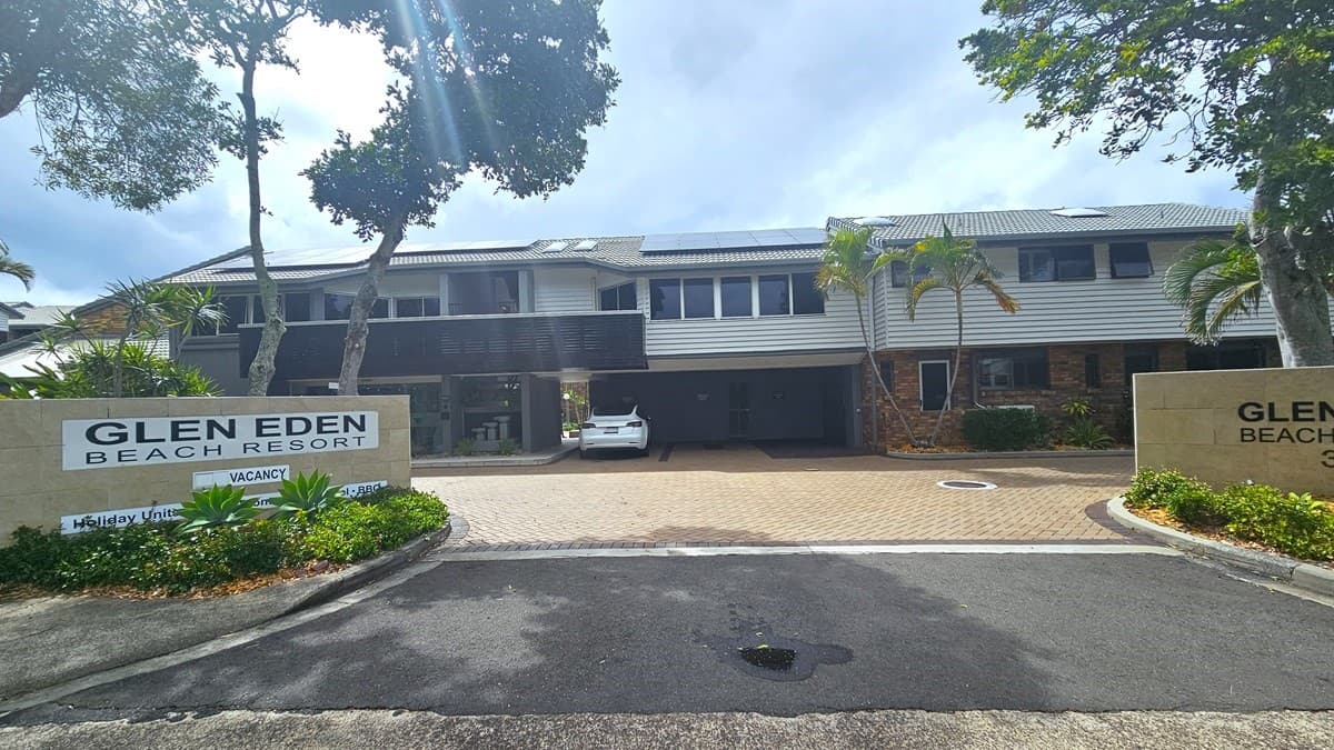 The main road is marked with white lines. On the right side, there is a two-storey building adorned with brown and white hues, highlighted by timber elements, and topped with grey corrugated iron roofing. The surrounding area is paved with brick pavers. Signage is positioned on both sides of the entrance, accompanied by landscaped plants and trees. Additionally, a white vehicle is parked in the undercover parking area to the right of the entrance.