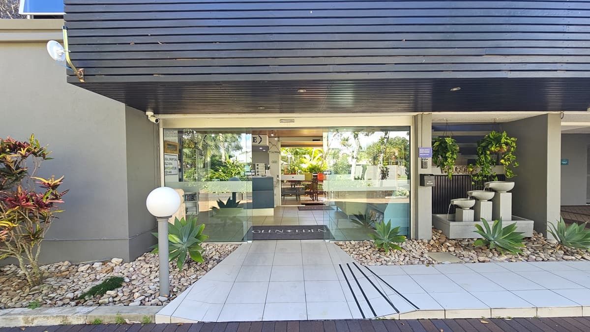 The facility entrance showcases an automatic glass sliding door with the facility logo. A grey mat lies on the doorstep. The concourse is paved with brick and transitioned to white tiles. A fountain, hanging ornamental plants and a landscape decorate the front. Black timber decorates the ceiling, while the walls are grey. A step-free ramp provides access to the entrance.