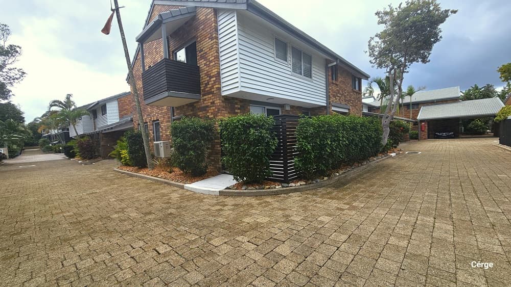 The driveway curves at the corner. In the corner stands a two-storey building with brown bricks and corrugated metal roofs. A metal fence and trimmed shrubs border the road. The driveway has brick designs. A landscape of plants and palm trees borders the road on the left.
