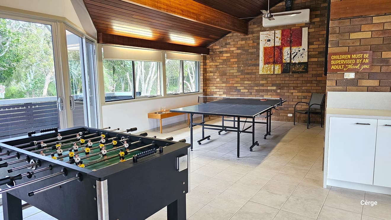 To the left, a foosball table and a table tennis setup. A bench chair is placed against the glass window, with an additional chair in the corner. A glass door leads to the balcony. To the right, there's a small kitchen area with a safety reminder on the wall. The ceiling boasts brown timber and a ceiling fan, while the walls display brown bricks. The floor is tiled in white. Abstract paintings decorate the brick wall.