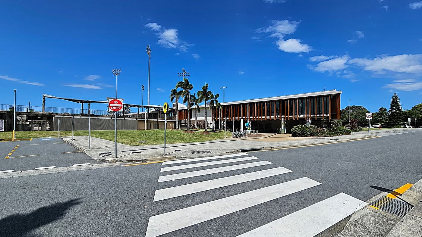 A modern building with vertical wooden slats on the right, surrounded by landscaped greenery. In front, a paved area with a bicycle rack and a few bicycles. Several palm trees are planted nearby. On the left, a sports field with tall floodlights and a "No Entry" sign. A zebra crossing is in the foreground, leading to a road with a "20" speed limit sign. The sky is clear with a few clouds.