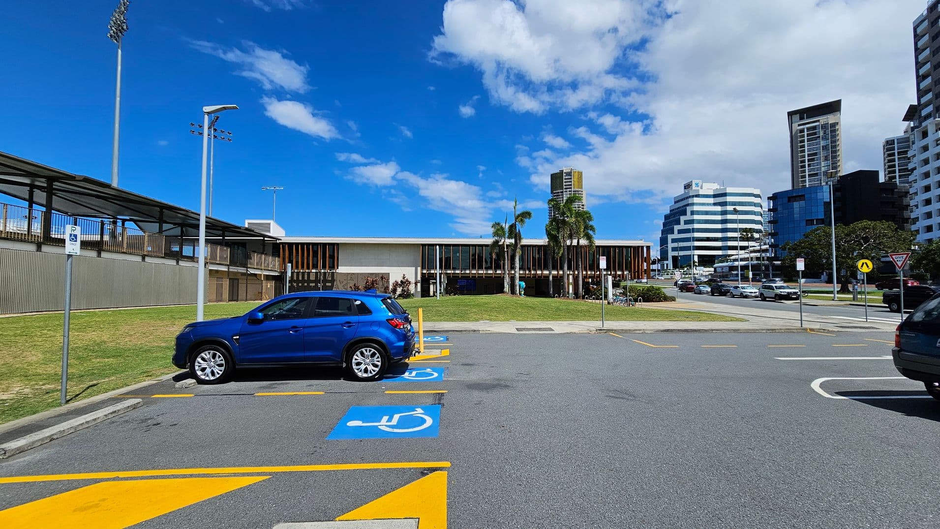 A car park with a blue vehicle in a designated disabled parking space on the left. The ground is marked with blue and yellow lines. In the background, a modern building with vertical wooden slats and palm trees in front. To the right, a road with several cars and tall buildings, including a distinctive white, tiered structure. A covered walkway and tall floodlights are visible on the far left.