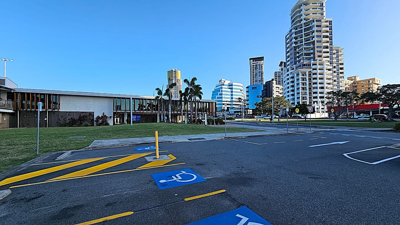 A parking area with marked disabled spaces in the foreground. On the left, there's a low, modern building with large windows and palm trees in front. In the background, several tall buildings with varied architectural styles rise against a clear sky. The ground is a mix of asphalt and grass, with a few road signs visible near the buildings.