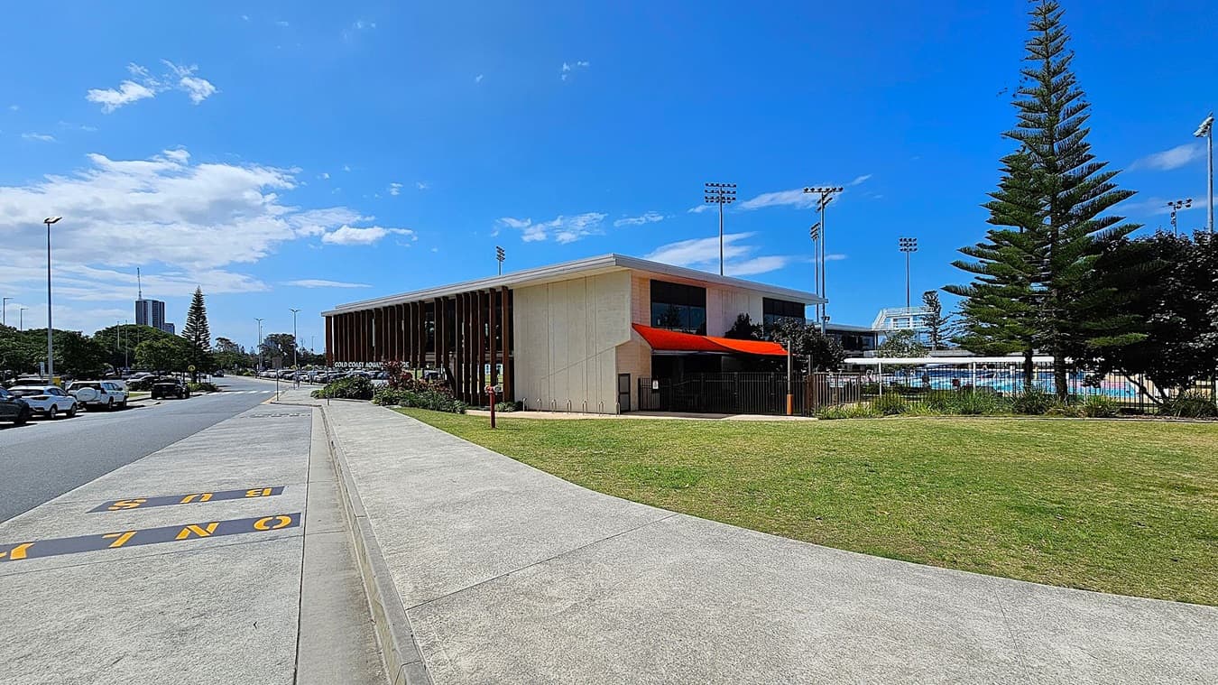 A modern building with a red awning is on the right, surrounded by a grassy area and tall trees. In the background, there are sports fields with floodlights. On the left, a road lined with parked cars and a "BUS ONLY" lane is visible. The pavement runs alongside the road, leading towards distant buildings and more greenery.