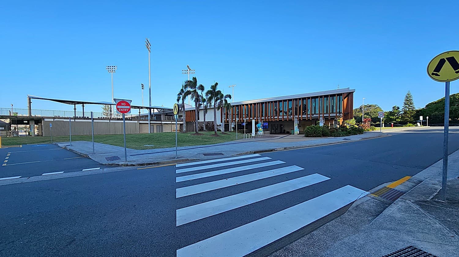 A street scene with a zebra crossing in the foreground. On the right, a yellow pedestrian crossing sign is visible. In the background, a modern building with large windows and wooden accents is surrounded by palm trees and greenery. A "No Entry" sign is on the left near a fenced area with tall floodlights. The road is clear, and the sky is bright blue.
