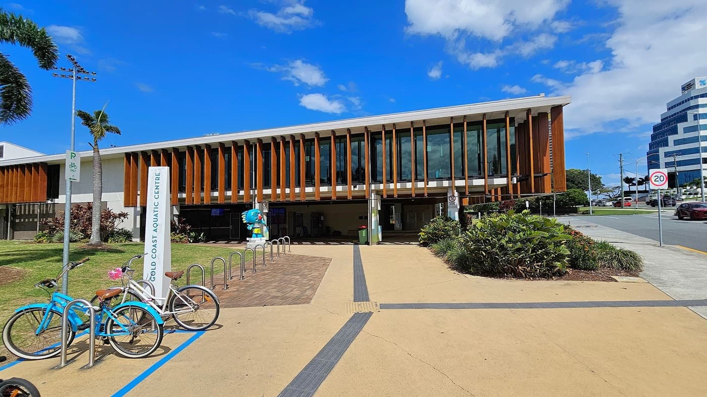A modern building with vertical wooden slats and large glass windows. In front, there's a sign reading "Gold Coast Aquatic Centre" and a bike rack with two bicycles. A colourful statue stands nearby. The foreground features a paved path with a textured strip. On the right, a road with a 20 km/h speed limit sign and a few cars. A palm tree and some greenery are on the left, with a tall building visible in the background.