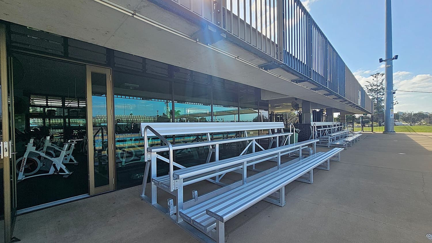 Outdoor area with a concrete floor and a metal railing above. On the right, there is a glass door leading to an indoor space with exercise bikes visible inside. In front of the glass door, there are metal bleachers. On the left, a lifeguard chair and a shaded area with a canopy are visible. In the background, there are more structures and a tall light pole.