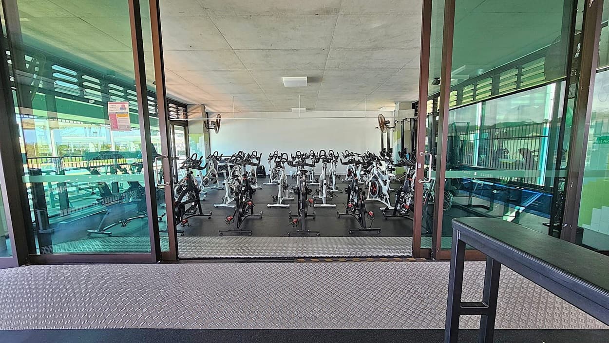A gym room with multiple stationary bikes arranged in rows on a dark floor. The room is enclosed by large glass doors, reflecting the outside. The ceiling is plain and light-coloured. In the foreground, there is a textured metal floor and a black bench on the right. The background shows a white wall and some gym equipment.