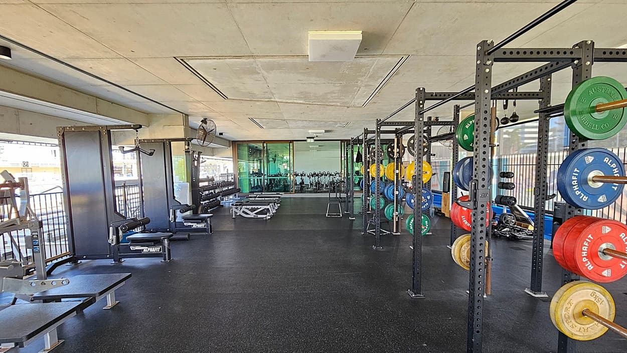 A gym with a black rubber floor and a concrete ceiling. On the left, there are weight benches and exercise machines near large windows. On the right, there are racks with colourful weight plates in green, yellow, blue, and red. In the background, more gym equipment is visible, including stationary bikes. The space is well-lit with natural light coming through the windows.